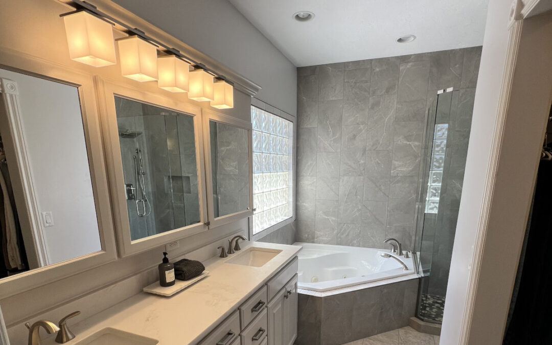 A modern, spacious bathroom featuring a white double vanity with rectangular mirrors, warm square lighting fixtures, and a corner jetted bathtub surrounded by large grey marble-look tiles