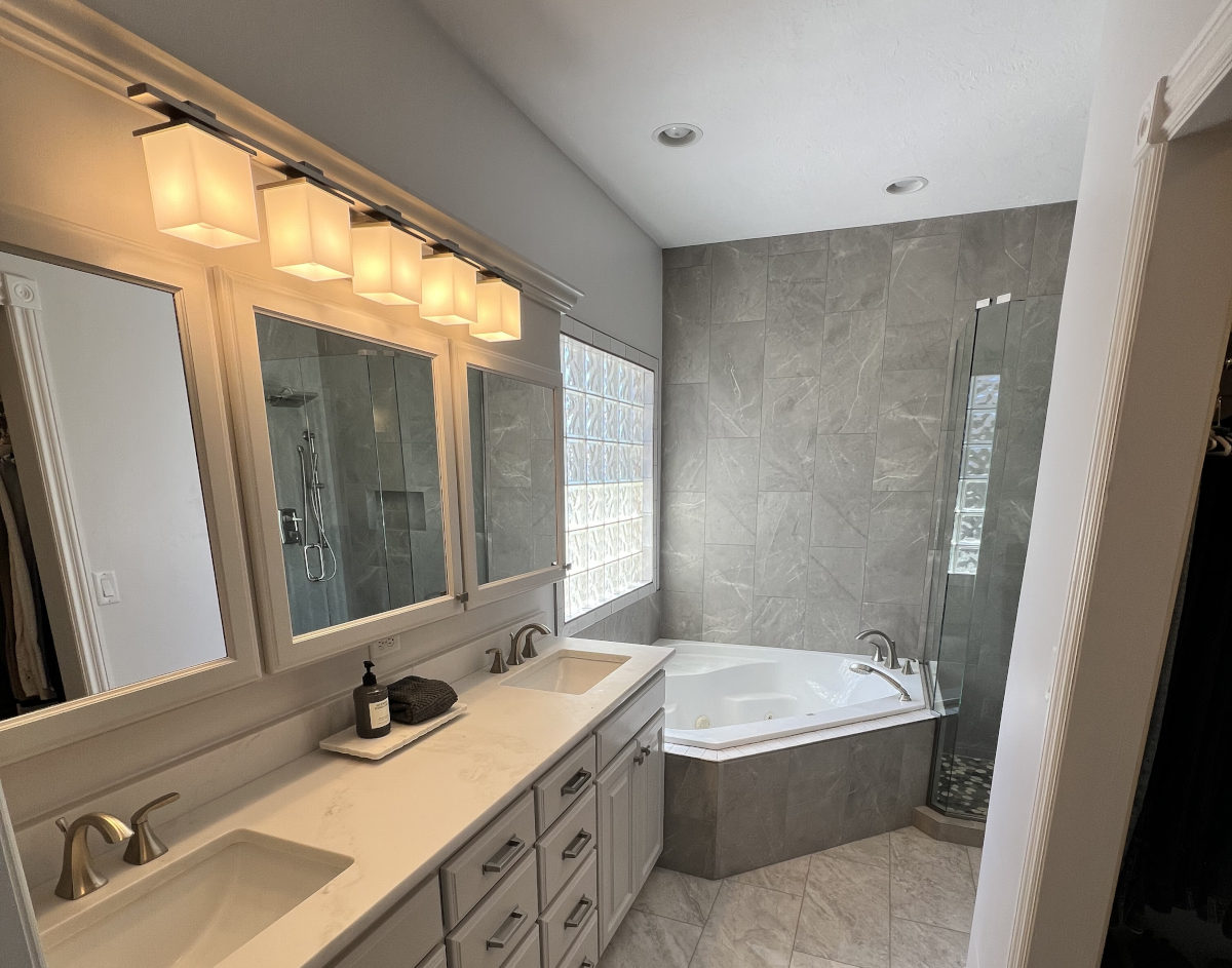 A modern, spacious bathroom featuring a white double vanity with rectangular mirrors, warm square lighting fixtures, and a corner jetted bathtub surrounded by large grey marble-look tiles